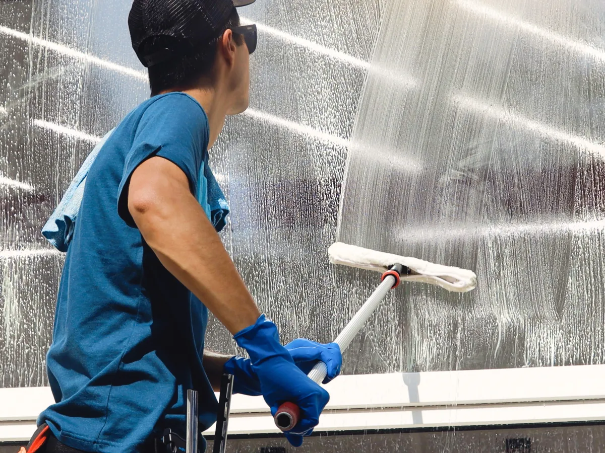 Cleaner in blue gloves washing a large window with a soapy applicator
